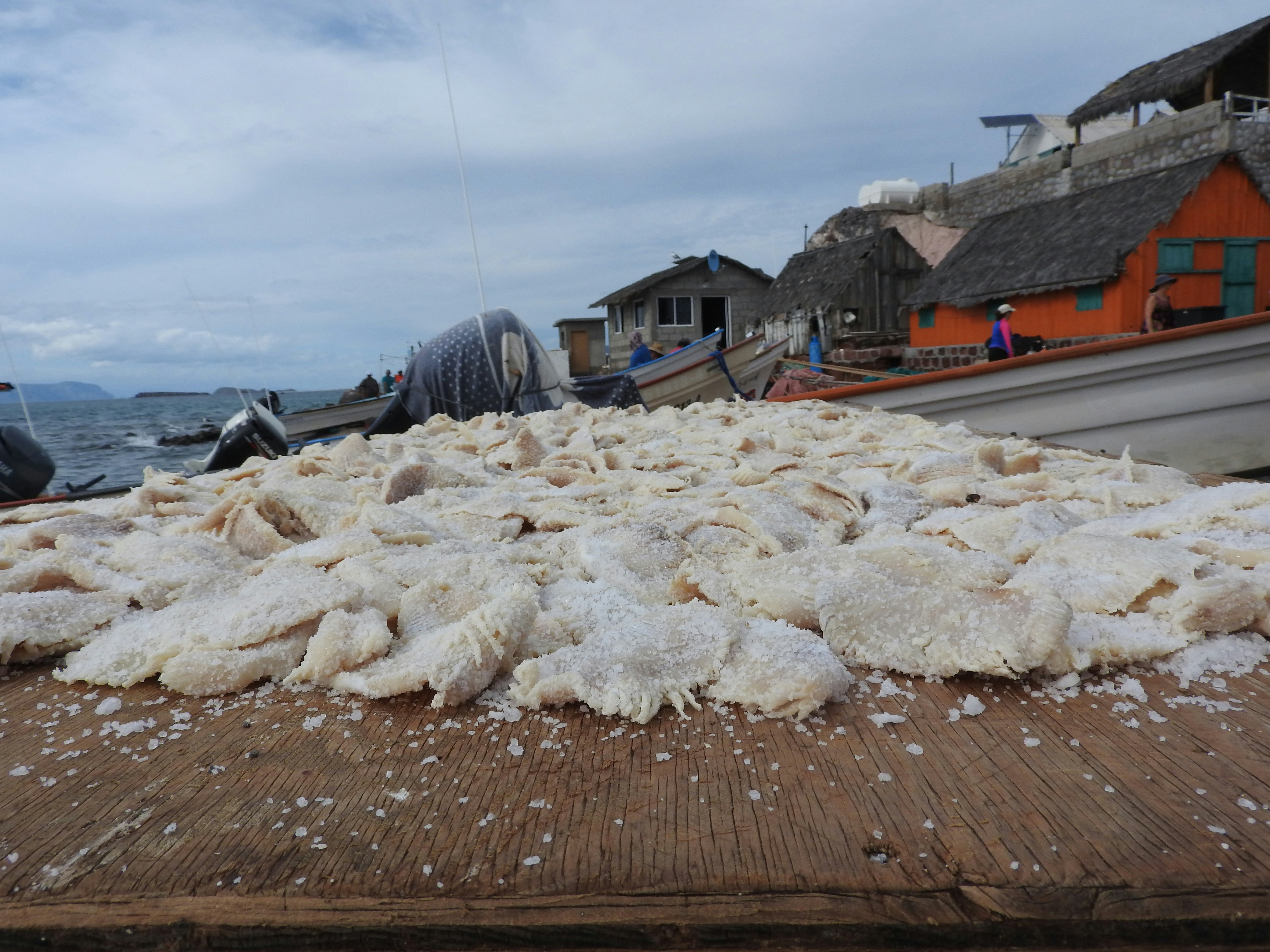 Coarse salt heaps spread across a wooden dock with boats and a colorful coastal village in the background.