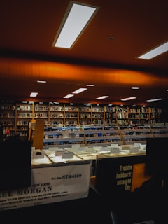 A dimly lit room with orange and warm lighting illuminates rows of shelves filled with books and records. The shelves are organized and labeled, and at the front of the image, several vinyl record bins are visible, each with labels for different artists. Fluorescent lights are embedded in the ceiling, contributing to the subdued ambiance.