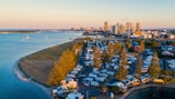 Aerial view of a coastal campsite with numerous RVs and caravans lined up next to a waterway, leading towards a city skyline in the distance. The area is bordered by a sandy beach and several tall trees, with calm water and a few boats visible. The cityscape includes modern high-rise buildings and appears to be near sunset, with warm lighting.