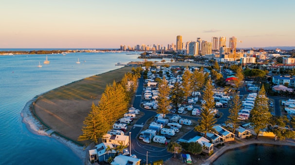 Aerial view of a coastal campsite with numerous RVs and caravans lined up next to a waterway, leading towards a city skyline in the distance. The area is bordered by a sandy beach and several tall trees, with calm water and a few boats visible. The cityscape includes modern high-rise buildings and appears to be near sunset, with warm lighting.