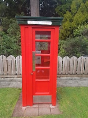 A bright red vintage telephone booth with a green roof and a sign displaying 'TOLL LOCAL CALLS TELEGRAMS'. It stands on a concrete path and is surrounded by a wooden fence and lush green plants in the background.