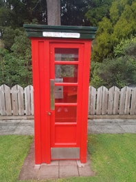 A bright red vintage telephone booth with a green roof and a sign displaying 'TOLL LOCAL CALLS TELEGRAMS'. It stands on a concrete path and is surrounded by a wooden fence and lush green plants in the background.