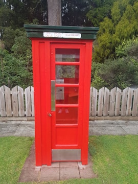 A bright red vintage telephone booth with a green roof and a sign displaying 'TOLL LOCAL CALLS TELEGRAMS'. It stands on a concrete path and is surrounded by a wooden fence and lush green plants in the background.