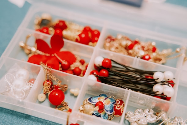 A plastic organizer with compartments holding various jewelry and hair accessories. The items include red and white beads, gold chains, and colorful enamel pieces. Some compartments contain decorative pins and earrings.