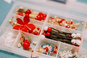 A plastic organizer with compartments holding various jewelry and hair accessories. The items include red and white beads, gold chains, and colorful enamel pieces. Some compartments contain decorative pins and earrings.