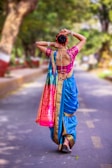 A woman is seen from behind, walking down a tree-lined street. She is wearing a traditional colorful saree with a turquoise blue base, pink and gold patterns. She has adorned her hair with flowers and is holding it up in a bun. The scene is bright and appears to be set outdoors on a sunny day.