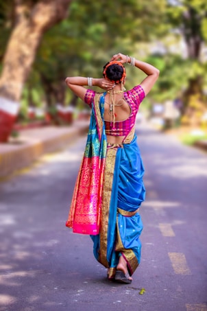 A woman is seen from behind, walking down a tree-lined street. She is wearing a traditional colorful saree with a turquoise blue base, pink and gold patterns. She has adorned her hair with flowers and is holding it up in a bun. The scene is bright and appears to be set outdoors on a sunny day.