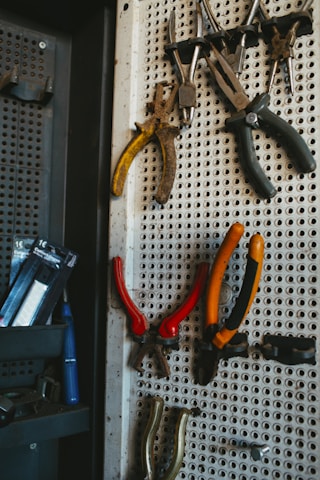 A selection of various hand tools, including pliers and wire cutters, hanging on a perforated metal pegboard. The tools have handles in different colors such as yellow, black, red, and orange. There are additional tools or items placed on a shelf to the left side of the image.