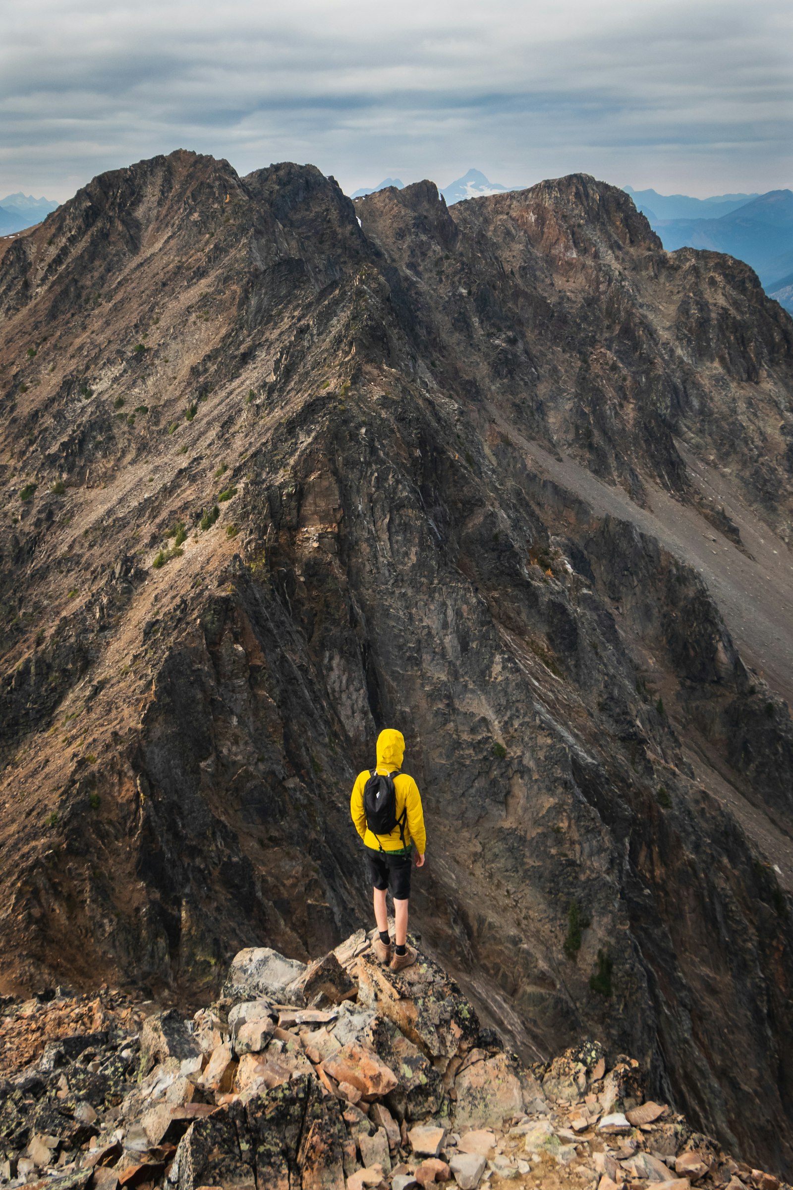 Hiker in a yellow shell jacket standing on a rocky ridge, snow-capped peaks beyond