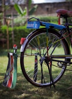 A colorful bicycle with a custom design featuring bright colors and personalized text. The back wheel has vibrant decorations and signage that reads 'Mark 1' and 'Unique Shuttle'. The bicycle stands on a grassy surface, with a blurred green natural background.