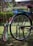 A colorful bicycle with a custom design featuring bright colors and personalized text. The back wheel has vibrant decorations and signage that reads 'Mark 1' and 'Unique Shuttle'. The bicycle stands on a grassy surface, with a blurred green natural background.