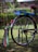 A colorful bicycle with a custom design featuring bright colors and personalized text. The back wheel has vibrant decorations and signage that reads 'Mark 1' and 'Unique Shuttle'. The bicycle stands on a grassy surface, with a blurred green natural background.