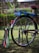 A colorful bicycle with a custom design featuring bright colors and personalized text. The back wheel has vibrant decorations and signage that reads 'Mark 1' and 'Unique Shuttle'. The bicycle stands on a grassy surface, with a blurred green natural background.