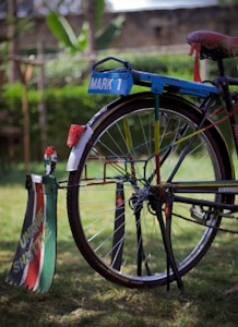 A colorful bicycle with a custom design featuring bright colors and personalized text. The back wheel has vibrant decorations and signage that reads 'Mark 1' and 'Unique Shuttle'. The bicycle stands on a grassy surface, with a blurred green natural background.