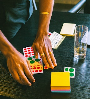 Hands are organizing various colored stickers and papers on a dark wooden table. A glass with some liquid residue stands nearby, along with stacks of colorful sticky notes.