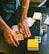 Hands are organizing various colored stickers and papers on a dark wooden table. A glass with some liquid residue stands nearby, along with stacks of colorful sticky notes.