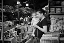 A woman holding a small child is standing in front of a market stall, which is filled with various ceramic bowls, plates, and other kitchenware. The woman is looking at something in her hand, possibly a phone, while the child looks on curiously. The scene is set in a busy outdoor market environment.