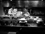 A monochrome image of two people working behind a food counter in a restaurant kitchen. The man, wearing a white chef's outfit, is preparing food, while the woman, in a striped shirt and apron, appears to be assisting. Pots and pans are arranged on the counter with small signs indicating various dishes like crispy pancake and fried pork spring roll. The background shows a busy kitchen with utensils and cooking equipment.