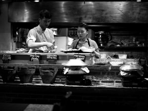 A monochrome image of two people working behind a food counter in a restaurant kitchen. The man, wearing a white chef's outfit, is preparing food, while the woman, in a striped shirt and apron, appears to be assisting. Pots and pans are arranged on the counter with small signs indicating various dishes like crispy pancake and fried pork spring roll. The background shows a busy kitchen with utensils and cooking equipment.