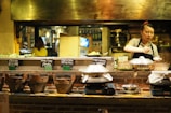 A woman prepares food in a rustic, warmly-lit kitchen. Various ingredients and dishes are displayed on a wooden countertop, with signs indicating items like crispy pancakes and fried wonton with crabmeat. Traditional clay stoves and bowls are prominently featured, creating an inviting, artisanal atmosphere.