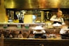 A woman prepares food in a rustic, warmly-lit kitchen. Various ingredients and dishes are displayed on a wooden countertop, with signs indicating items like crispy pancakes and fried wonton with crabmeat. Traditional clay stoves and bowls are prominently featured, creating an inviting, artisanal atmosphere.