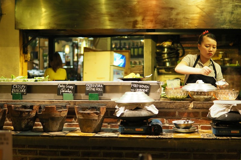 A woman prepares food in a rustic, warmly-lit kitchen. Various ingredients and dishes are displayed on a wooden countertop, with signs indicating items like crispy pancakes and fried wonton with crabmeat. Traditional clay stoves and bowls are prominently featured, creating an inviting, artisanal atmosphere.