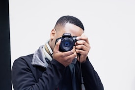 A person holds a DSLR camera up to their face, capturing a photograph. They wear a black jacket and a multi-colored scarf. The background is plain and minimal, creating focus on the subject.