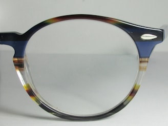 A close-up view of a pair of eyeglasses with a dark-colored, semi-transparent frame. The frame features subtle mixes of blue, brown, and clear hues. The lens is clear, and there is a small metallic ornament near the hinge.
