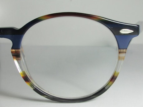A close-up view of a pair of eyeglasses with a dark-colored, semi-transparent frame. The frame features subtle mixes of blue, brown, and clear hues. The lens is clear, and there is a small metallic ornament near the hinge.