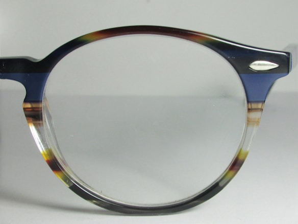 A close-up view of a pair of eyeglasses with a dark-colored, semi-transparent frame. The frame features subtle mixes of blue, brown, and clear hues. The lens is clear, and there is a small metallic ornament near the hinge.