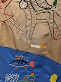 Colorful embroidery depicting abstract shapes, a chair, a cigarette with smoke, a fish, and other designs on a fabric background divided into brown and blue sections.