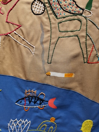 Colorful embroidery depicting abstract shapes, a chair, a cigarette with smoke, a fish, and other designs on a fabric background divided into brown and blue sections.