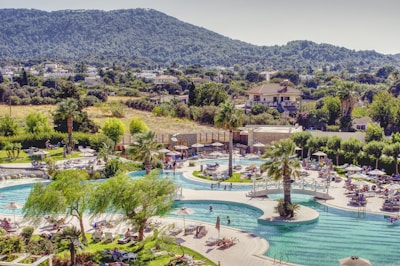 A resort pool area with people relaxing on sun loungers under umbrellas. The pool is large with bridges and surrounded by palm trees and various green plants. In the background, there are hills covered in dense greenery and a few houses scattered in a rural setting.
