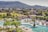 A resort pool area with people relaxing on sun loungers under umbrellas. The pool is large with bridges and surrounded by palm trees and various green plants. In the background, there are hills covered in dense greenery and a few houses scattered in a rural setting.