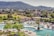 A resort pool area with people relaxing on sun loungers under umbrellas. The pool is large with bridges and surrounded by palm trees and various green plants. In the background, there are hills covered in dense greenery and a few houses scattered in a rural setting.