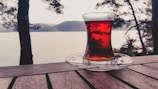 A glass of tea rests on a wooden table with a saucer beneath it. In the background, there are silhouettes of trees and a serene body of water with hills on the horizon.