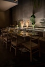 A dimly lit dining area with a long table set for a meal. The table is adorned with glassware, napkins, and decorative dried plants as centerpieces. Wooden chairs surround the table, and the backdrop features dark walls decorated with indoor plants, adding a natural touch to the setting.