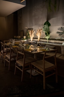 A dimly lit dining area with a long table set for a meal. The table is adorned with glassware, napkins, and decorative dried plants as centerpieces. Wooden chairs surround the table, and the backdrop features dark walls decorated with indoor plants, adding a natural touch to the setting.