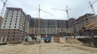 A large construction site with a multi-story building framework. Several cranes are positioned around the structure, indicating active construction. Scaffolding and construction materials are visible around the area, and a truck is parked near the building.