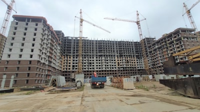 A large construction site with a multi-story building framework. Several cranes are positioned around the structure, indicating active construction. Scaffolding and construction materials are visible around the area, and a truck is parked near the building.