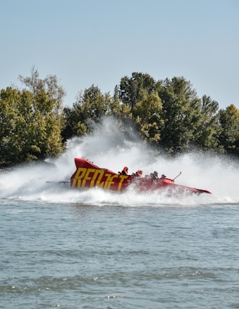 A red speedboat with the text 'RED JET' on its side races across a body of water, creating large splashes. Several people wearing helmets are on board, enjoying the high-speed ride. The background is lush with green trees under a clear blue sky.