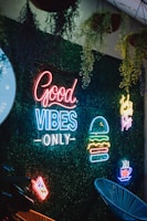A vibrant neon sign display against a backdrop of green foliage. The signs include phrases like 'Good Vibes Only' and images of a burger, a steaming cup, and a slice of pizza. Hanging plants are visible above, adding a natural element to the scene.