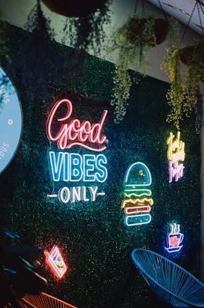 A vibrant neon sign display against a backdrop of green foliage. The signs include phrases like 'Good Vibes Only' and images of a burger, a steaming cup, and a slice of pizza. Hanging plants are visible above, adding a natural element to the scene.