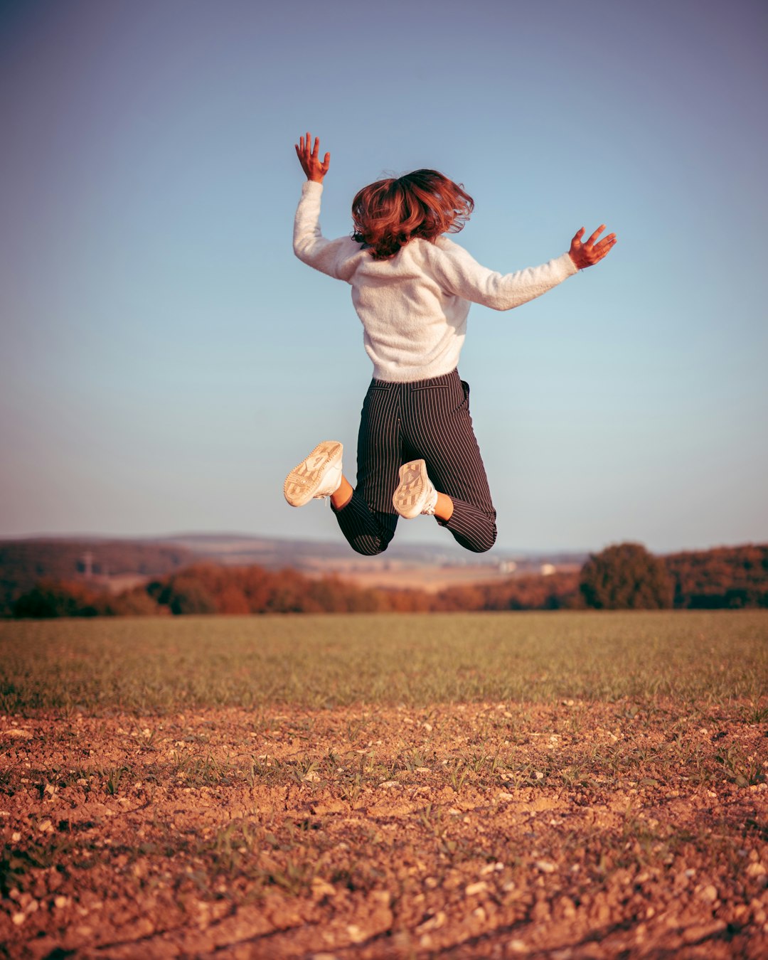 woman in white long sleeve shirt and black pants jumping on brown field during daytime