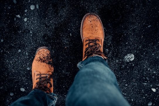 A close-up view of a person's feet wearing brown leather boots, standing on a dark, rocky surface. The boots have small bits of debris on them, suggesting a rugged or outdoor environment. A pair of jeans is visible as part of the attire.