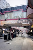 A bustling outdoor market with various clothing stalls, located beneath buildings with prominent signage. Clothes are on display both hanging on racks and spread across tables. There is visible debris and uneven pavement, possibly indicating ongoing construction or recent damage. People are walking through the market, with one person in the foreground wearing a red and black checkered coat and looking back at the camera.