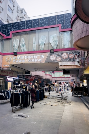 A bustling outdoor market with various clothing stalls, located beneath buildings with prominent signage. Clothes are on display both hanging on racks and spread across tables. There is visible debris and uneven pavement, possibly indicating ongoing construction or recent damage. People are walking through the market, with one person in the foreground wearing a red and black checkered coat and looking back at the camera.