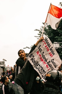A group of people gathered in a protest. One individual is prominently holding a sign with Indonesian text and a red-and-white Indonesian flag. The crowd appears to be animated and engaged, with some wearing face masks and others making gestures. A police helmet is visible in the crowd.