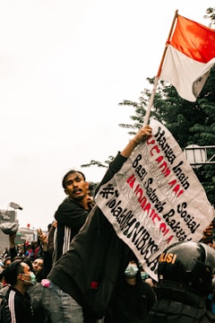 A group of people gathered in a protest. One individual is prominently holding a sign with Indonesian text and a red-and-white Indonesian flag. The crowd appears to be animated and engaged, with some wearing face masks and others making gestures. A police helmet is visible in the crowd.