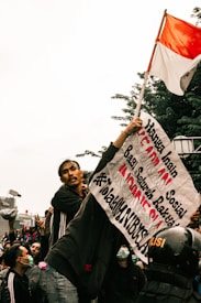 A group of people gathered in a protest. One individual is prominently holding a sign with Indonesian text and a red-and-white Indonesian flag. The crowd appears to be animated and engaged, with some wearing face masks and others making gestures. A police helmet is visible in the crowd.